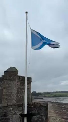 The video shows the Scottish flag waving atop a white pole next to a stone castle under a cloudy sky. The castle has a round tower and multiple windows. Beyond the castle, the landscape reveals a flat, open area with distant green hills. The camera slowly pans upward to capture the vast expanse of the cloudy sky above.