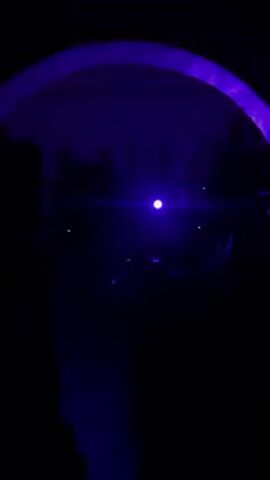 The scene is a dimly lit bar, illuminated by a strong blue spotlight and purple ambient lighting. The camera is positioned behind an archway, through which the bar area is visible. A man in a white t-shirt walks into the foreground. A man without a shirt is visible in the background, near the bar. Shelves holding bottles are visible behind the men. A speaker is visible on the left side of the frame. The overall atmosphere suggests a dance or club setting.