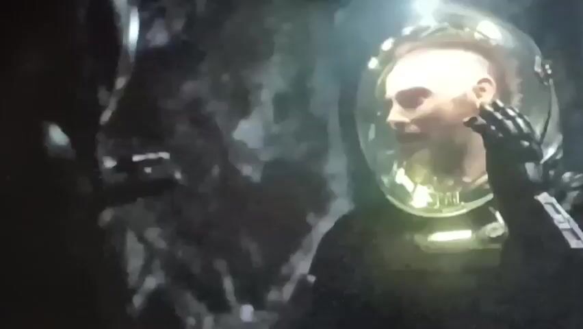 A man wearing a plaid blanket over his shoulders is looking down at a holographic display. The display shows a complex network of lines and shapes in blue. In the background, there are screens with green graphics, one of which appears to be a medical scan. To the right of the main display, a man in a diving suit with a clear helmet is visible. He is looking forward and his face is illuminated from within the helmet. In the background, there are rocky, cavern-like structures.