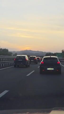 The video shows a view from a car on a highway with several other cars in front. The sky is a light orange and yellow, suggesting it is either sunrise or sunset. Mountains are visible in the distance. The cars are moving in a line on the highway, and the overall scene depicts a traffic situation with a scenic background.