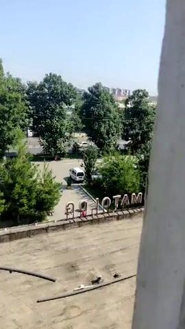 The video shows a high-angle view looking down at a street scene with trees and buildings. In the foreground, there is a concrete ledge with pipes running along it. Below the ledge, there is a sign with large, metallic letters spelling out "RADIOTAM". A white van is parked in a lot behind the sign, and several trees line the street. Further in the background, there are buildings and a clear blue sky. The scene appears to be outdoors during the daytime.