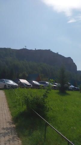 This video shows a scenic view of a grassy area with a mountain in the background. Several cars are parked in a row next to the grass. Small structures, possibly gazebos or cabins, are scattered throughout the grassy area. The sky is blue with some scattered clouds, suggesting it's a clear day. The sun is high in the sky, indicating it is likely daytime, possibly mid-morning or afternoon. The overall mood is peaceful and serene, with the natural landscape dominating the scene. There is no visible text in the video. The setting appears to be a natural park or recreational area, possibly a campsite or a tourist destination, given the presence of parked cars and the structures. The location context of Yuldybaevo, Russia, suggests a rural or natural environment.