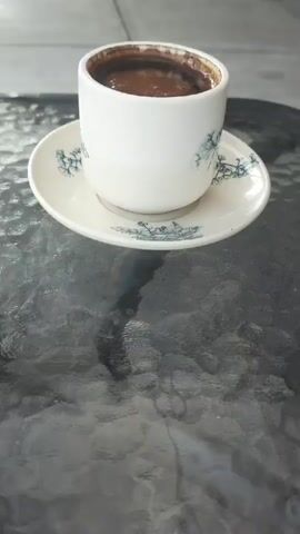 A close-up shot focuses on a white ceramic cup filled with dark liquid, likely coffee, resting on a matching saucer. The cup and saucer have a delicate blue pattern. The items are placed on a textured glass surface, possibly a table. In the blurred background, outdoor seating areas with tables and chairs are visible, suggesting a cafe or restaurant setting. The overall lighting appears natural, indicating it might be daytime.