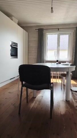 A video shows a kitchen area with white cabinets and an oven on the left. A white round table with chairs is positioned in the center of the room, in front of a window with gray curtains. The floor is made of wooden planks. A black chair with metal legs is in the foreground, facing the table and window. The lighting suggests it is daytime.