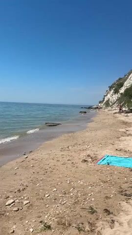 The video shows a wide sandy beach under a clear blue sky. The gentle waves of the turquoise sea lap against the shore. In the distance, a rocky cliff covered in green vegetation rises from the coastline. A few people can be seen walking along the beach and near the cliff, some possibly swimming or relaxing. A bright blue beach towel is laid out on the sand in the foreground, and a blue umbrella is visible further down the beach. The scene is peaceful and appears to be a pleasant day at the beach.
