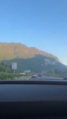 A vehicle travels down a highway in the daytime. In the distance, a large mountain is visible. The car’s dashboard is visible with orange stitching. A train track runs parallel to the highway. Other cars are visible on the road. A rearview mirror shows the mountain and train track.
