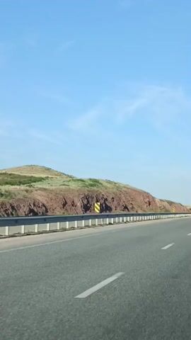 This is a vertical, first-person perspective video taken from a moving vehicle. The road is asphalt with white lane markings and a metal guardrail on the left. To the left of the guardrail is a rocky, reddish-brown hillside with sparse green vegetation. The sky is clear and blue with a few wispy clouds.