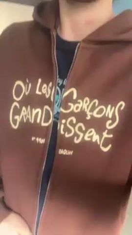 A person is zipping up a brown zippered hoodie. The hoodie has the words “Où Les Garçons GRANDissent” in white lettering across the chest, along with the text “N° 9901 EAQLHV” beneath it. The person has a beard and there is a red mark visible on their neck. A blue T-shirt can be seen underneath the open hoodie. The background consists of light blue walls and a glimpse of a white door.