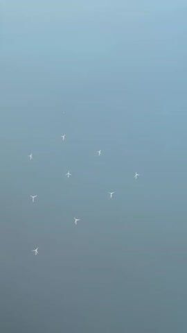 An aerial shot shows a cluster of white wind turbines scattered across a pale blue expanse. The turbines are evenly spaced and appear to be located on the water, suggesting an offshore wind farm. The perspective is from high above, looking down on the turbines and the hazy sky. There are no people or other significant objects visible in the scene. The lighting is soft, indicating it might be early morning or late afternoon.