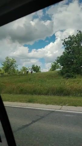 The video shows a view from inside a vehicle looking out the window. The landscape is green with rolling hills covered in grass and trees. The sky is blue with scattered white clouds. There are several people visible in the distance on one of the hills. They appear to be walking or standing. The road is visible in the foreground. The video is shaky, suggesting it was taken while the vehicle was in motion.