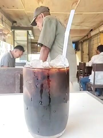 The video depicts a glass of dark liquid, possibly coffee or soda, filled with ice and a white straw. The glass sits on a white table in what appears to be a small, open-air cafe or restaurant. Behind the glass, an elderly man wearing a cap and a khaki shirt is standing and interacting with something out of frame. In the background, two other men are seated at tables. One is leaning over a table, seemingly engaged in an activity, while the other sits upright. The setting appears to be a casual, local eatery with a simple, perhaps rustic, interior.