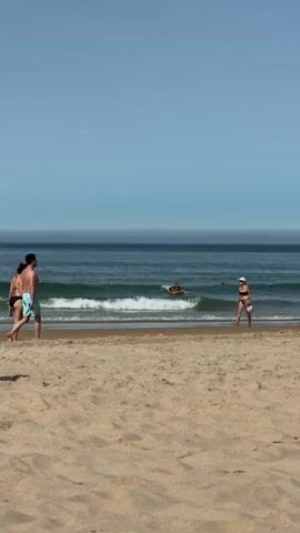 A video shows a sandy beach with people interacting. In the foreground, the sand is smooth. In the mid-ground, a group of four people are walking together near the water's edge. Two of them are women wearing bikinis, and two are men wearing swim trunks. Further out in the ocean, one person is on a surfboard. To the right, another person in a white hat and dark bikini walks along the water. In the background, the ocean stretches out under a clear blue sky.