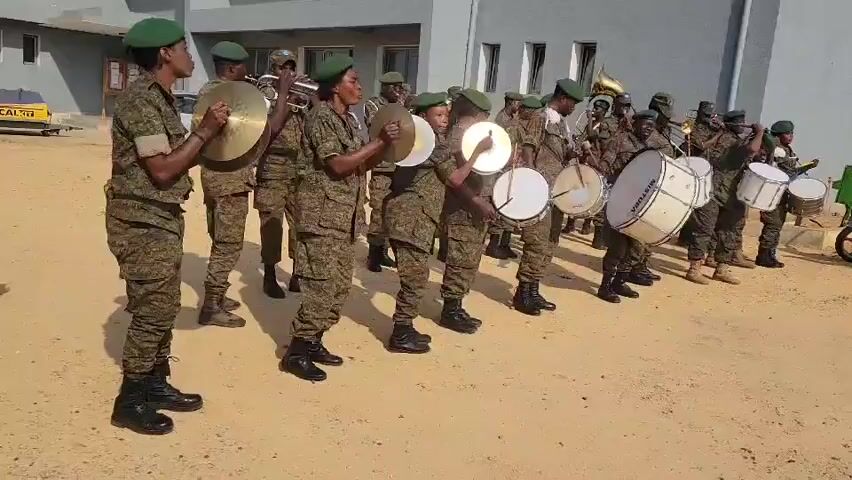 A military band composed of approximately fifteen individuals, all wearing camouflage uniforms and green berets, stands in formation on a dirt surface in front of a grey building. The band members are playing various instruments, including drums and cymbals. The individuals in the foreground are playing cymbals, while those further back are playing drums and brass instruments. The lighting suggests it is daytime.