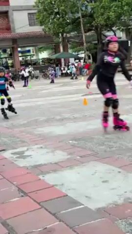 Several children wearing roller skates are participating in a training session or activity course. They are skating around small, orange cones set up in a large, paved area that looks like a public square or outdoor space. All the children are wearing helmets and knee pads, and some have elbow pads as well. They are dressed in colorful athletic wear. The pavement is made up of large, off-white tiles interspersed with rows of red bricks. Behind the skating area is a building with covered walkways, trees, and parked vehicles. Some adults are standing around watching the children. The camera is positioned near the ground, possibly on a curb or low wall, and remains mostly static.