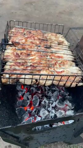A close-up shot shows chicken pieces being grilled on a barbecue. The chicken is arranged in rows on a metal grill basket, which is positioned over a bed of glowing hot coals. The chicken appears to be seasoned and is beginning to brown and char in places. The grill is set in a black metal barbecue pit. The background is out of focus but appears to be an outdoor setting with dirt or gravel on the ground. The video is a continuous shot of the grilling process, with subtle camera movements and zooms. There are no people visible in the frame.