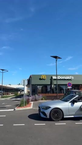 The video shows a silver convertible driving past a McDonald's restaurant. The car is moving from left to right in the frame. A person is visible in the driver's seat of the car, holding a brown paper bag. The McDonald's restaurant has green siding and the iconic golden arches logo. The sky is blue with some clouds. There are lampposts and other cars visible in the background. The video appears to be filmed outdoors during the daytime.