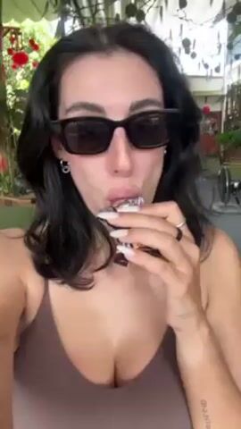 A woman wearing sunglasses and a brown tank top is eating a candy bar. She is sitting in what appears to be an outdoor restaurant or cafe with an informal seating area and ceiling fans. The woman looks directly at the camera, takes a bite, and seems to be enjoying her treat. The background includes plants, chairs, and glimpses of a bar area.