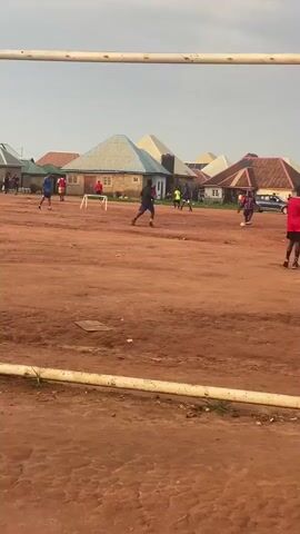 A group of young men are playing soccer on a dirt field. Several houses with colorful roofs are visible in the background. One man in a red shirt and black shorts kicks the ball, while another man in a dark shirt and shorts runs to intercept it. A soccer goal with white posts stands on the right side of the frame. The sky is cloudy.