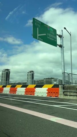 The video shows a view from inside a car moving on a road, likely a bridge or highway. The road has wet patches and painted white lines, and is flanked by orange and white safety barriers on one side and a grey concrete barrier topped with a wire fence on the other. Through the fence, several tall, modern apartment buildings are visible against a partly cloudy sky. Road signs are present, including one in Korean. The reflection of the car's interior and the sky can be seen in the windshield.