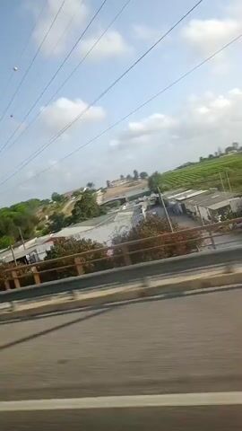 The video is a wide shot from a moving vehicle, looking out over a landscape. The foreground consists of a highway with a concrete barrier. Beyond the barrier, there are several buildings with metal roofs, some trees, and a large green field. In the distance, there are more trees and a few buildings on a hill. The sky is blue with some scattered clouds. There is a billboard with the Mitsubishi Electric logo visible on the right side of the frame. The video appears to be taken during the daytime.