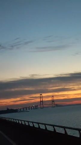 The video shows a view of a long suspension bridge over water at sunset. The sky is a mix of dark blue at the top and orange near the horizon, with some clouds scattered across the sky. The bridge has tall towers and lights along its length. The camera is likely moving on another bridge or roadway, as a railing is visible in the foreground. The water is calm, reflecting some of the sky's colors.