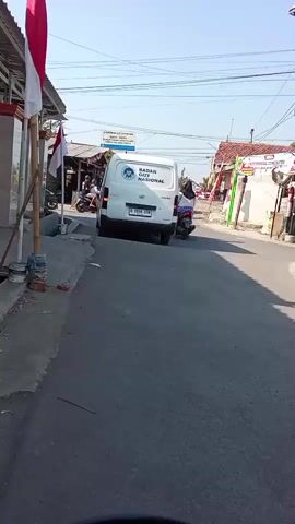 A white van with text in Indonesian visible on its back travels down a paved road. Indonesian flags are visible on the left side of the road. A person on a scooter rides behind the van.