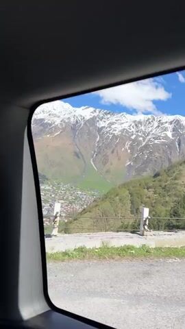 The video shows a view of a snow-capped mountain range and a town nestled in a valley, taken from a moving vehicle. The mountains are the main focus, with patches of snow visible against the rocky terrain. Below the mountains, a town with buildings and green fields stretches out. The viewpoint is from a car window, with a roadside barrier visible in the foreground. As the video progresses, the view shifts to a close-up of trees passing by the window, intermittently revealing glimpses of the mountains in the background. The sky is blue with scattered clouds.