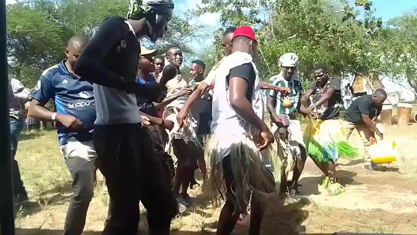 Here is a description of the video.

The video shows a group of men dancing outdoors in a grassy area. Some of the dancers are wearing traditional attire, including grass skirts and headwraps. Other men wear t-shirts, jeans, and caps. Several tents and trees are in the background, and the sky is bright. One man appears to be gathering yellow balloons. The dancers seem to be enjoying themselves. The music playing sounds like a mix of African and electronic beats.