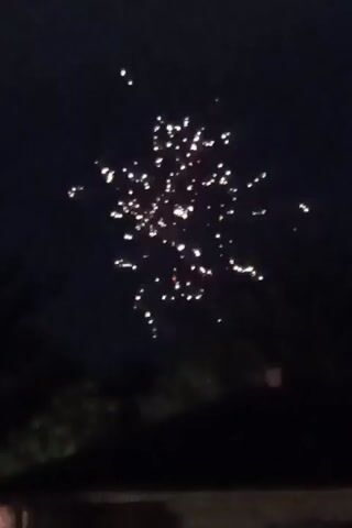 The video displays a night sky with several small, illuminated objects. At the beginning of the video, there is a cluster of these lights. As the video progresses, some lights appear to move or ascend. One frame shows a brief burst of light resembling a firework. Other lights are visible scattered across the sky, with some appearing to be stationary while others move. The scene appears to be outdoors, with dark foliage and the silhouette of a roofline visible at the bottom of the frame. The lighting conditions are dark, suggesting it is nighttime.
