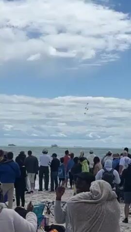 Here is a forensic description of the video.

The video captures a crowd of people gathered on a sandy beach, gazing upwards at a formation of four fighter jets soaring across a partly cloudy, blue sky. The aircraft, likely part of an aerial display team, maintains a tight, diamond-shaped formation. The beachgoers are a mix of ages and ethnicities, dressed in casual attire suitable for a beach outing, and seem captivated by the aerial display. Some are pointing or holding up their phones, probably to record the event. The sea is visible in the background, with at least two large vessels in the distance, suggesting the event is taking place near a busy maritime route. The overall atmosphere is one of awe and excitement as the crowd witnesses the aerial spectacle.