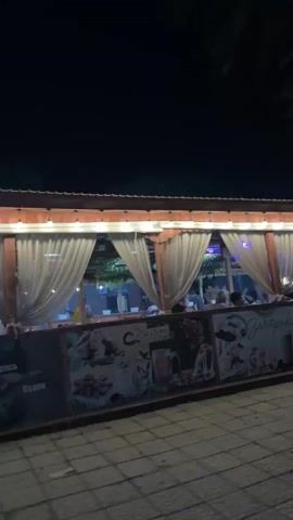 A nighttime exterior shot of a restaurant's covered seating area. The area is enclosed by white curtains and decorated with string lights. People are seated inside, dining. A tiled walkway is visible in the foreground. The overall atmosphere is dimly lit and appears to be a casual dining establishment.