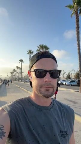 A man wearing a dark grey sleeveless shirt, a black baseball cap, and sunglasses is filming himself with a phone. He is walking or jogging on a paved pathway alongside a beach. The sun is low in the sky, casting a bright glow and creating lens flare. Palm trees line the pathway, and in the background, there are cars parked, other people walking, and a Ferris wheel. Another person, a Black man wearing a red shirt and black shorts, is jogging on the pathway in the background. The overall environment is a beachside promenade on a sunny day.
