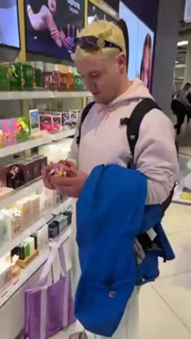 A young man with short, bleached blond hair, wearing sunglasses, a pink hooded sweatshirt with lettering, a backpack, and holding a bright blue jacket, is standing in a store aisle with shelves filled with perfume and cosmetics. He's examining a small, flower-shaped perfume bottle. The store is well-lit, with other shoppers visible in the background.