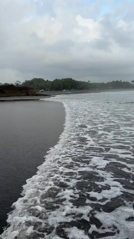The video shows a black sand beach on a cloudy day. Waves with white foam are coming into the shore on the right side of the frame. The sand is smooth and dark, contrasting with the foamy water. In the distance, there is a line of trees and vegetation along the coast, blending into the cloudy sky. The overall atmosphere is serene and somewhat overcast.