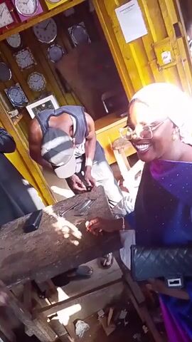 The video shows a small group of people in what appears to be a shop or market. A woman wearing glasses and a white headscarf is smiling and laughing, seated at a wooden table. A man in a gray tank top and baseball cap is standing at the table, seemingly working on something with his hands. Clocks are visible on the wall behind them. The camera shifts, showing another person in a patterned shirt, looking towards a man in a black jacket who is standing in the background. Overall, the setting appears somewhat crowded and the lighting is bright.