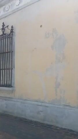 The video begins focused on a window with a decorative metal grate, set into a pale yellow wall with architectural details. The camera then pans to the left, revealing a narrow street lined with buildings painted in various colors including green, pink, and blue. Plants decorate some of the storefronts. A person walks in the distance. 

The camera continues to pan, showing a paved public square with trees, benches, and street lamps.  A building with a red tile roof and the inscription "Sala Actual Legislativo" is visible in the center of the square. Another building with white columns can be seen to the left. The sky is blue and clear. A lone person walks across the square.