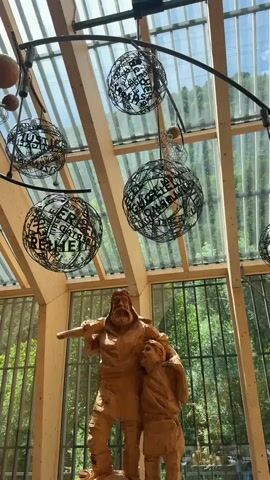 A wooden sculpture of a bearded man and a child stands beneath a modern glass and wood atrium. Hanging from the ceiling are several spherical metal sculptures adorned with text. The atrium has large windows that look out onto a green, wooded hillside. The lighting suggests it is daytime.
