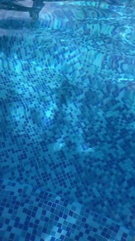 The video shows a close-up view of water in a swimming pool. The bottom of the pool is tiled with small blue and light blue square tiles. The water is clear and slightly rippling, causing distortions in the appearance of the tiles below. The camera appears to be held above the water, providing a direct, overhead perspective.