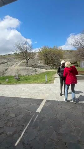 The video shows a paved outdoor area under a metal awning, with people gathered around. A Georgian flag is visible waving in the wind, adjacent to a parking area. Beyond the people, there is a stone path, grass, bare trees, and a rocky hillside under a blue sky with white clouds. Some people are standing and talking, while others appear to be waiting in line. The location seems to be a tourist site or a place of interest.