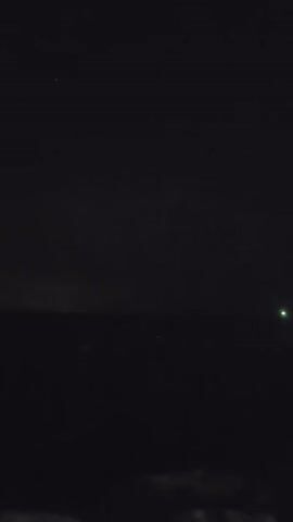 The video shows a night sky over a body of water. Lightning flashes intermittently in the distance, illuminating dark clouds. The water is choppy, and the scene is mostly dark, with some faint lights visible on the horizon in the later frames. The camera appears to be stationary, possibly handheld.