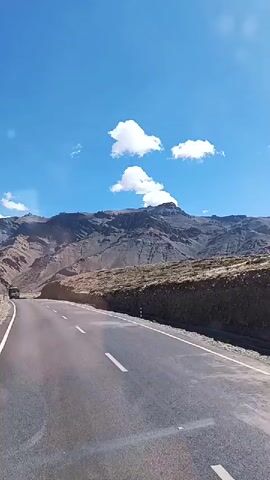 The video captures a roadside view of a mountainous landscape under a clear blue sky with scattered white clouds. A paved road curves from the left side of the frame and extends towards the background. On the right side of the road, a rough, rocky embankment rises steeply, covered with dry vegetation. In the distance, imposing, rugged mountains with rocky slopes dominate the horizon. A truck is visible on the road, moving away from the viewer and towards the mountains. The overall scene suggests a journey through a remote, high-altitude region.