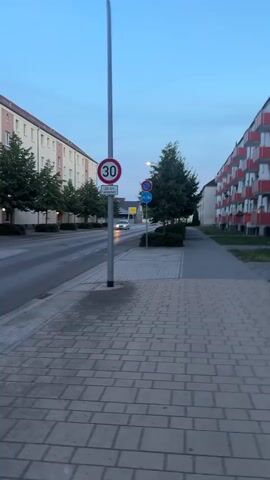 This is a vertical video taken from a sidewalk in Rathenow, Germany. The scene depicts a street with apartment buildings on both sides. On the left, a light-colored apartment building runs along the street, with trees lining the sidewalk in front of it. On the right, a white apartment building with red balconies stretches down the street.

The street itself is paved and appears to be a two-lane road with a white car visible driving in the distance. The sidewalk in the foreground is also paved with rectangular bricks.

Several traffic signs are visible. A speed limit sign indicating "30" is prominently displayed on a pole near the center of the frame. Below it, another sign with text, possibly indicating specific times or conditions for the speed limit, is partially visible. Further down the street, a blue circular sign with a red border and a diagonal red bar is visible, which could be a "no stopping" or "no parking" sign.

The time of day appears to be dusk or early evening, as the sky is a deep blue with some clouds. There is a slight haze in the air. The overall atmosphere is calm and quiet, suggesting a residential area. No people are clearly visible in the video, though there might be figures on some of the balconies of the building on the right. The video seems to be a static shot with some slight camera movement or stabilization.