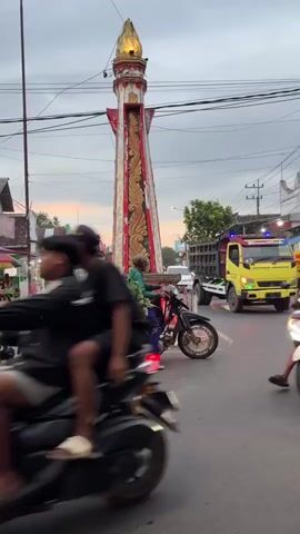 This video captures a bustling street scene in Kebonsari, Indonesia, likely during the late afternoon or early evening as suggested by the soft, diffused light and the warm hues of the sunset visible in the sky. The central focus is a tall, ornate monument, a distinctive landmark with red and gold decorative elements, topped with a golden flame-like structure.

The street is active with various modes of transportation. Several motorbikes are prominent. One motorbike carries a large load of green produce, likely bananas or similar, tied to the back, with a person seated behind the driver. Another motorbike has two young men riding it, appearing to be in motion. A third motorbike, with a person in the foreground, is also moving, with a blurred appearance due to its speed. Other motorbikes are visible, some appearing to be parked.

In the background, there are shops and buildings lining the street, some with awnings and signs that are difficult to read clearly. One sign on a building appears to have some text on it. There's a glimpse of a large truck in the distance, suggesting commercial activity.

The overall atmosphere is one of everyday life and commerce. People are engaged in their daily routines, transporting goods and traveling. The presence of the monument indicates a public space, possibly a town square or intersection. The weather appears to be clear, contributing to the visibility of the warm sky. The scene is vibrant and full of movement, showcasing the lively character of the location.