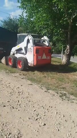A white Bobcat S175 skid steer loader with orange rims is parked on a gravelly path next to a dark green dump truck. The dump truck is positioned behind the skid steer loader. Trees and vegetation are visible in the background, along with a building to the left and a utility pole. The scene appears to be outdoors on a sunny day.