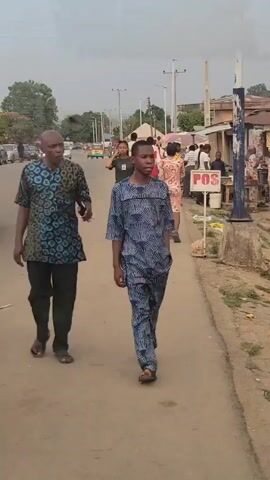Here is a description of the video:
The video shows a street in an urban area, likely in Nigeria, given the style of dress and signage. People are walking along the side of the road. Two men in matching patterned outfits walk towards the camera. Behind them, a small group of women are walking, and then a woman in a green t-shirt and black pants walks into view. Along the edge of the road is a small table with what looks like produce being sold, and a sign that says "POS". Other people and cars are visible in the background. The weather appears overcast.