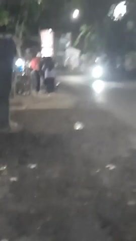 The video captures a dimly lit outdoor scene, possibly at dusk or night, with a focus on a road and its immediate surroundings. The ground appears to be a mixture of dirt and asphalt, with some debris scattered across it. In the background, a street or road is visible, with a vehicle's headlights shining brightly, indicating movement. On the left side of the frame, a group of people stands near what appears to be a cart or stall. One individual is wearing a red top, and others are in darker clothing. The overall ambiance is somewhat blurry and grainy, suggesting it was filmed in low-light conditions. The scene suggests a casual gathering or waiting by the roadside.