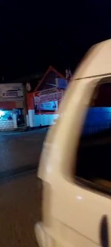 The video shows a street scene at night, decorated with red and white banners and flags, likely celebrating Indonesian Independence Day. There are several motorcycles parked on the left side of the street, and a few people are visible, some sitting and some riding motorcycles. The main banner reads "DIRGAHAYU RI KE-81," which translates to "Happy 81st Indonesian Independence Day." The environment appears to be an urban or semi-urban area with buildings on either side of the street. The lighting is artificial, from streetlights and building lights, creating shadows and highlighting the decorations. The camera is moving, suggesting someone is walking or driving through the area.