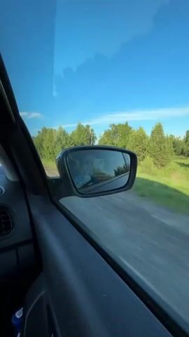 The video shows a view from inside a moving car, looking out the passenger side window and into the side mirror. The car is traveling on a paved road through a rural area with lush green trees and a bright blue sky. In the side mirror, a person is visible, presumably the driver, wearing sunglasses and holding a phone. The car's interior, including a black door panel, is also visible in the foreground. The motion blur of the passing scenery indicates the car is in motion.