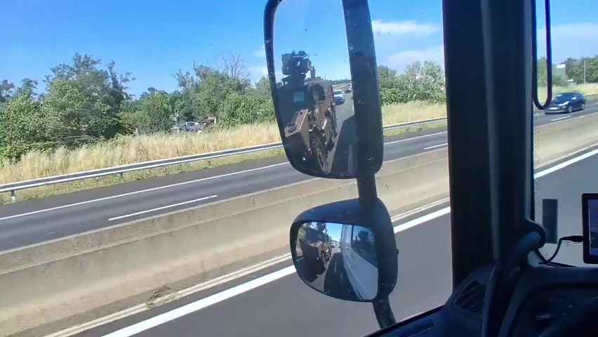 A first-person perspective shot from the cab of a truck shows a convoy moving along a highway. The truck is driving next to a large, brown military vehicle, possibly an armored personnel carrier. The side mirrors of the truck provide views of the road behind, showing other vehicles, including semi-trucks and cars. The highway has multiple lanes and a concrete barrier on the side. The weather appears sunny. Inside the truck cab, there are two GPS devices mounted on the dashboard displaying maps. The environment outside the truck includes green trees and some buildings in the distance.