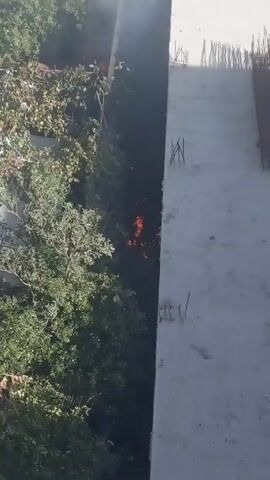 The video shows a fire burning in a narrow space between buildings, obscured by trees. Smoke rises from the flames, which are visible in the lower center of the frame. To the right, a concrete structure with exposed rebar is visible. Green trees and foliage are to the left. The camera is positioned above the scene.